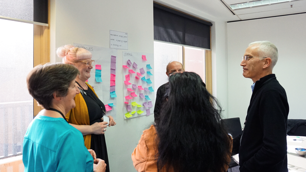 A group of five individuals in a meeting room, standing and facing a wall covered with colorful sticky notes. Their faces are obscured for privacy. There is a whiteboard on the wall with some text that is not legible from this angle. The room has large windows allowing natural light to enter, and there’s a closed door visible in the background